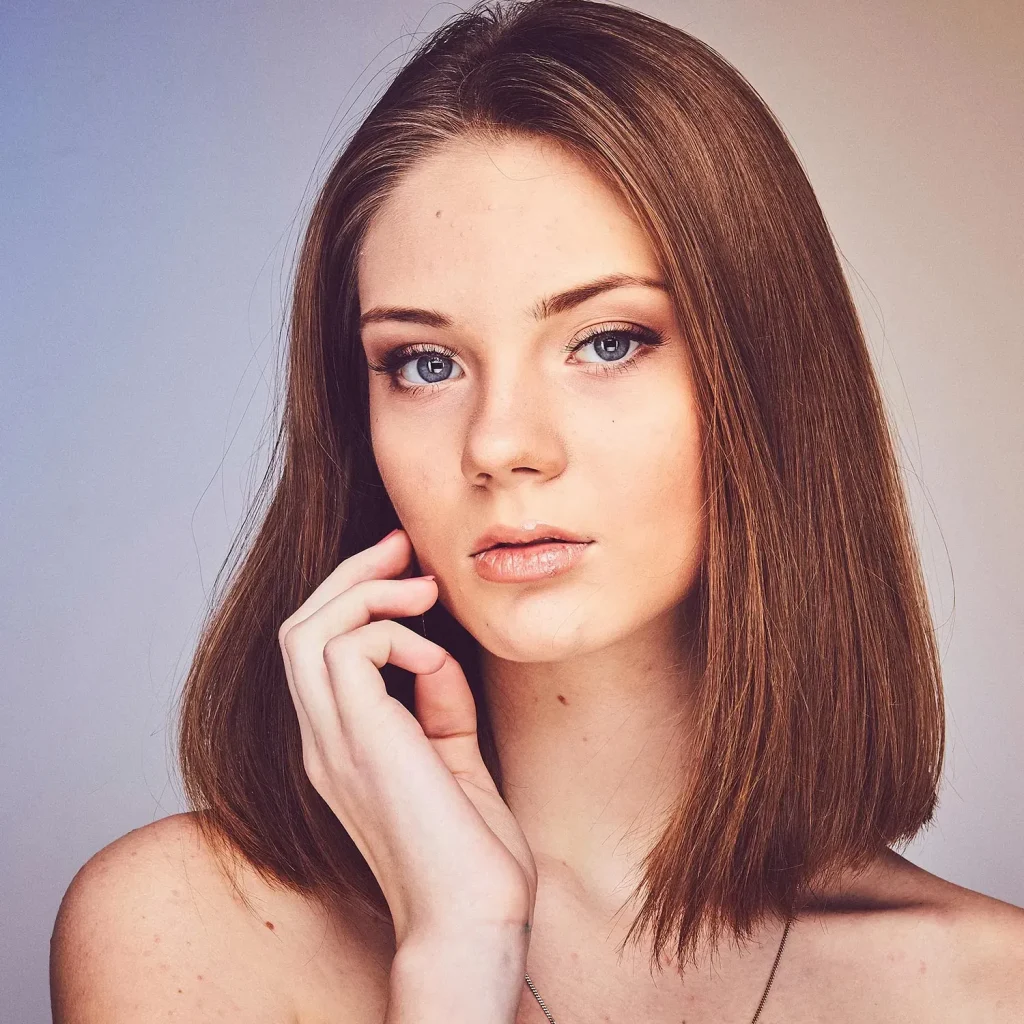 A young woman with straight, shoulder-length brown hair and blue eyes gazes at the camera, gently touching her face with one hand against a neutral background.
