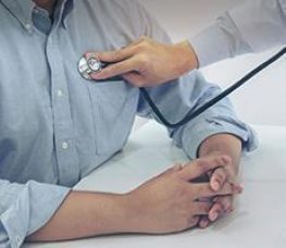 A healthcare professional uses a stethoscope to listen to a patients chest while the patient sits with hands folded on a white table.