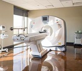 A modern CT scanner sits in a bright, clean medical room with polished floors and large windows, surrounded by medical equipment.