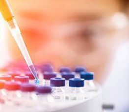 A close-up of a scientist using a pipette to place liquid into vials with blue caps, with the scientist’s face blurred in the background.