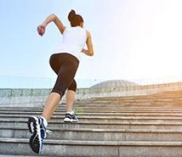 A woman in athletic wear runs up concrete stairs outdoors, with sunlight shining in the background.