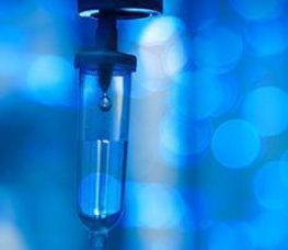Close-up of a medical IV drip chamber with a drop of fluid inside, set against a blurred blue background with circular light patterns.