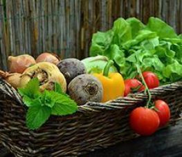 A wicker basket filled with fresh vegetables including tomatoes, onions, beets, a yellow bell pepper, lettuce, and a sprig of mint, set against a wooden background.