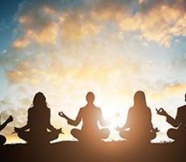 Five people sit cross-legged in a row, meditating with their hands in a mudra position, silhouetted against a bright sunrise or sunset sky with scattered clouds.
