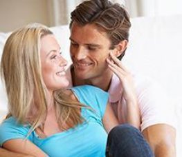 A smiling couple sits close together on a couch, looking into each other’s eyes. The woman has blonde hair and wears a blue shirt, while the man has brown hair and wears a light pink shirt.