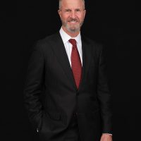 A middle-aged man with gray hair and a beard, wearing a black pinstripe suit, white shirt, and red tie, stands and smiles against a solid black background.