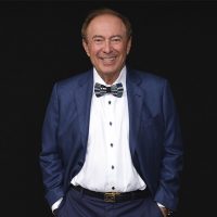A smiling man wearing a blue checkered suit, patterned bow tie, and white shirt stands with his hands in his pockets against a black background.