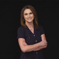 A woman with straight brown hair stands against a black background, smiling with her arms crossed. She wears a navy short-sleeve blouse with gold buttons and simple jewelry.