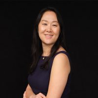 A woman with long dark hair, wearing a sleeveless navy dress, smiles and poses with arms crossed against a plain black background.